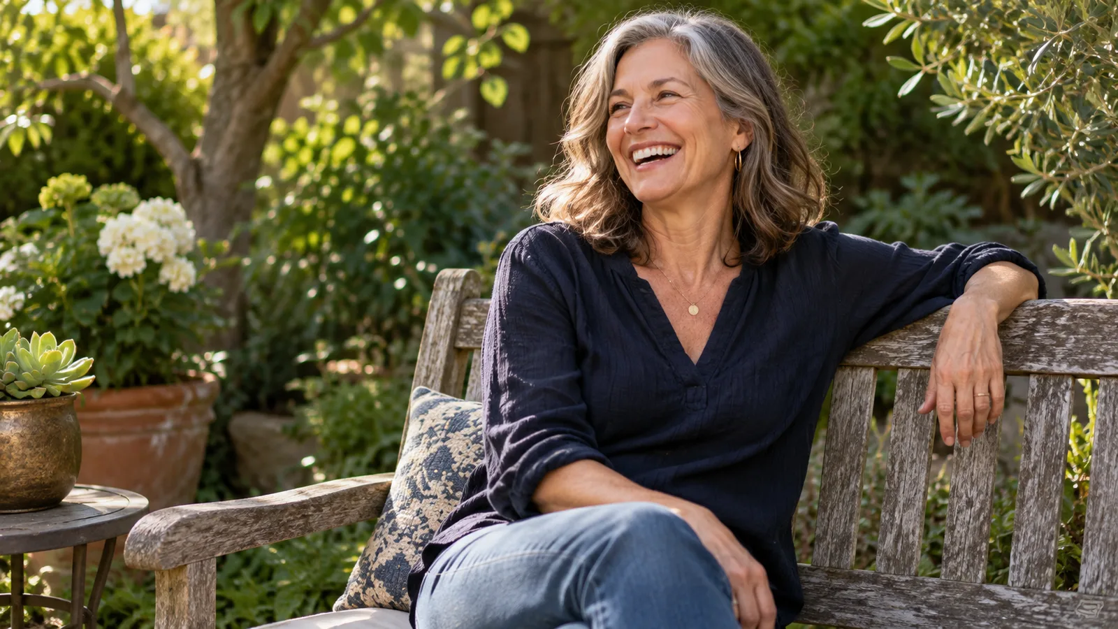 A smiling woman with grey hair relaxes on a wooden garden bench in sunlight